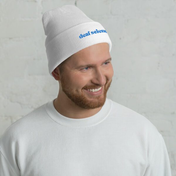 Person with short hair wearing white beanie with blue "deaf scientist" embroidery