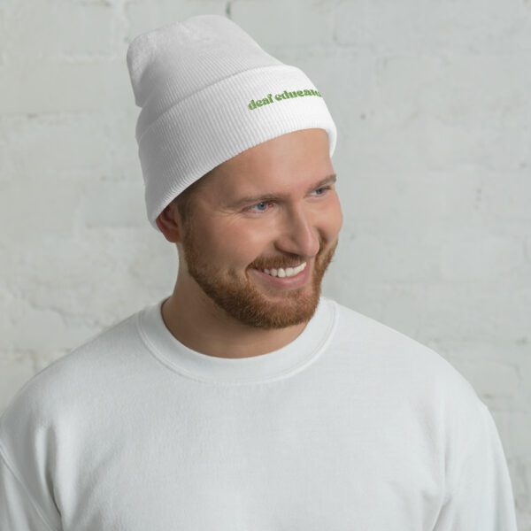 Person with short hair wearing white beanie with green "deaf educator" embroidery