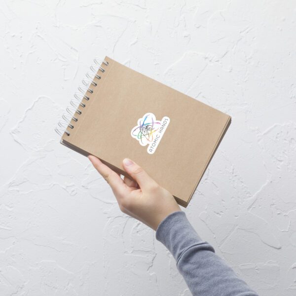 A hand holding a brown notebook with the Atomic Hands logo sticker in the middle